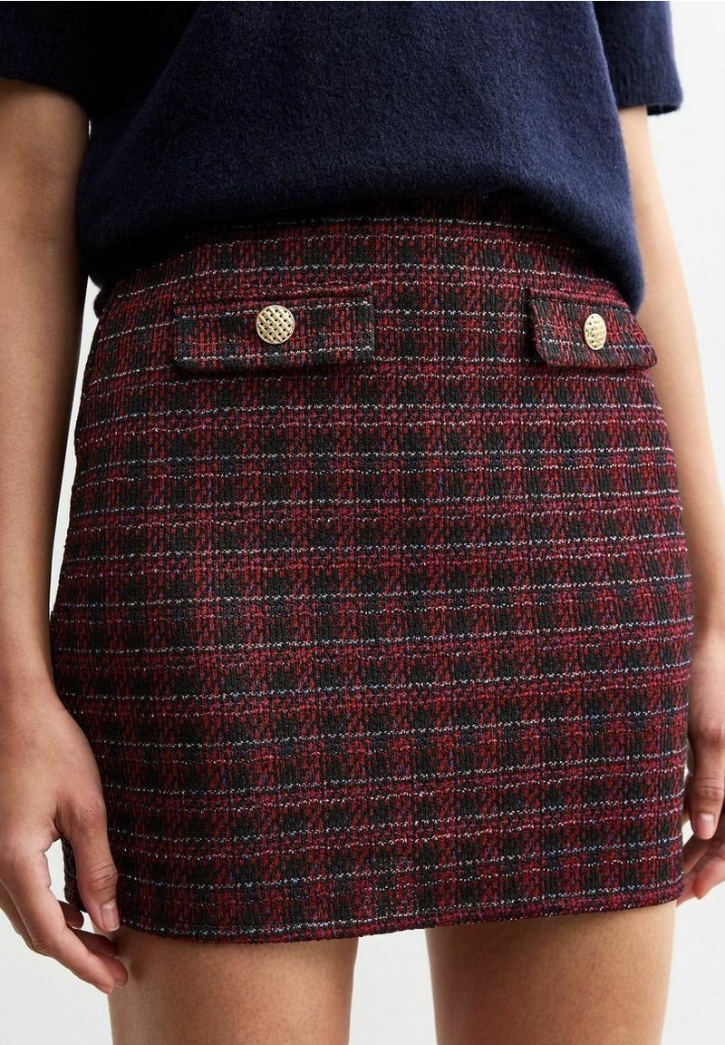 Gros plan d'une personne portant une mini-jupe en tweed rouge et noir à motifs avec deux poches à rabat à l'avant et des boutons dorés.
