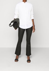 Persoon in witte trui over witte shirt, zwarte leren flare broek, zwarte hoge hakken sandals, houdt een zwarte tas in de linkerhand.