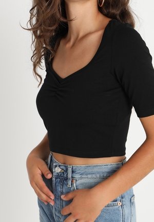 Frau mit welligen braunen Haaren, die goldene Creolen trägt, ein schwarzes kurzärmeliges Cropped-Top und hoch taillierte blaue Jeans, die Hände ruhen nahe der Taille.