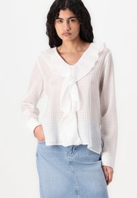 Blusa blanca texturizada con un cuello con volantes y cierre frontal. Manga larga con puños, combinada con una falda de mezclilla azul claro.
