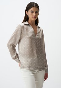 Camicia color taupe chiaro con scollo a V e colletto, caratterizzata da pois bianchi. Realizzata in un tessuto morbido e semi-trasparente con maniche lunghe.