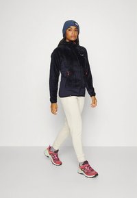 Tamsiai mėlyna vilnos striukė su pūkuotu audiniu, šoniniais kišenėmis ir kontrastinėmis oranžinėmis užtrauktukais, derinama su smėlio spalvos kelnėmis ir daugių spalvų sportiniais bateliais.