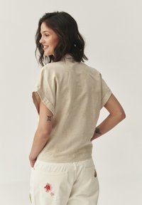 Camisa beige de manga corta con un patrón floral texturizado, que presenta un cuello tipo camiseta. Combinada con pantalones blancos que tienen bordados florales.