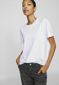Kvinne som har på seg en enkel hvit t-skjorte med korte ermer og svarte jeans, stående med hendene i lommene, mot en lys grå bakgrunn.