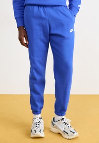 Kék sportnadrág elasztikus derékkal, felhúzható bokákkal és egy kis fehér Nike logóval. Simább anyagtextúra, világos sportcipőkkel párosítva.
