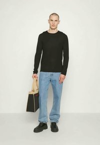 Cămașă lungă neagră cu mâneci, blugi drepți de culoare albastră deschisă, pantofi negri, ținând o geantă mică neagră cu litere roșii, pe un fundal neutru.