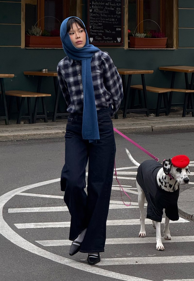 A person in a plaid shirt and blue scarf walks a Dalmatian in a black shirt and red beret on a pink leash. Urban background with empty tables.