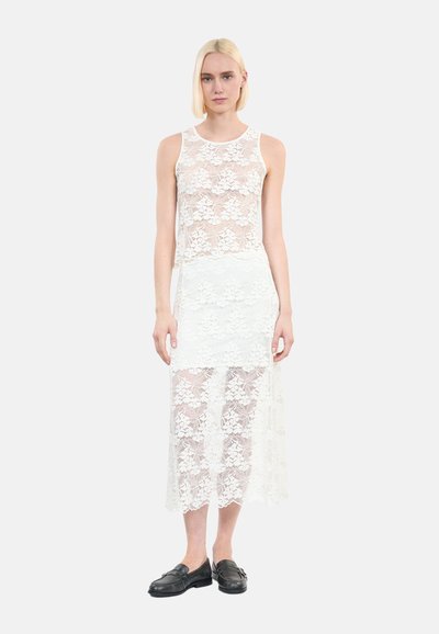 Robe blanche en dentelle avec un design superposé ; haut sans manches, jupe au genou, motifs floraux et texture semi-transparente. Chaussures noires portées.