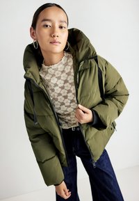 Giacca a vento verde con cappuccio, dotata di chiusura con zip, polsini elastici e dettagli neri, indossata sopra un top in pizzo e jeans scuri.