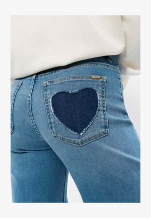 Vaqueros azul claro con un parche en forma de corazón azul oscuro en el bolsillo trasero, combinados con una camiseta blanca.