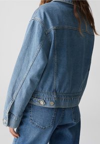 Džínová bunda ve světle modré barvě s uvolněným střihem, spadlými rameny, uzávěrem na dva knoflíky a detailem na zadním díle. Stehy jsou viditelné a povrch má texturovaný finiš.