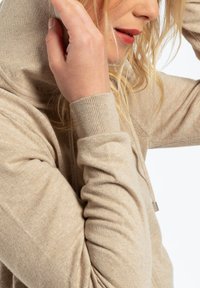 Haut en tricot beige avec col montant, poignets côtelés et tissu texturé. Comprend des cordons de serrage et un design décontracté.