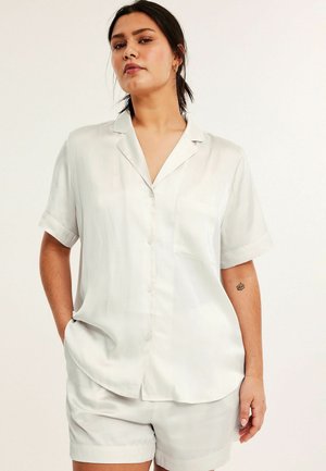 Kvinde med mørkt hår i en lav hestehale, iført en hvid kortærmet skjorte med knapper og matchende shorts, stående med den ene hånd i lommen.