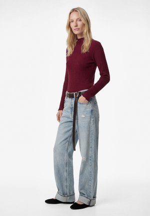 Kvinde stående iført en ribbet, burgunderfarvet langærmet top, lyseblå vidde-ben jeans med opbuk ved kanten og et langt brunt bælte samt sorte flade sko.