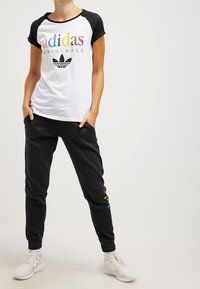 Osoba stojící s rukama v kapsách, oblečená v bílém tričku Adidas Originals s barevným nápisem a černých teplácích s bílými teniskami.