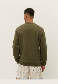 Olivgrüner Pullover mit rundem Halsausschnitt und gerippten Bündchen, aus weichem Stoff. Getragen mit gemusterten hellen Shorts mit floralen Designs.
