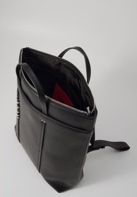 Zaino in pelle nera con apertura superiore con zip, tasca esterna e due spallacci regolabili. L'interno presenta un design con logo e un oggetto rosso.