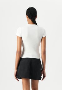 Top blanc à manches courtes en maille côtelée avec une coupe ajustée, porté avec une jupe noire comportant deux poches arrière. Texture lisse, silhouette simple.