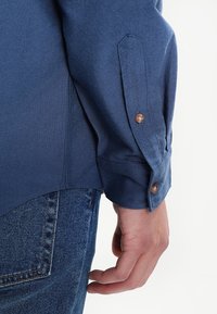 Blaue strukturierte Hemd mit gekrempelten Ärmeln und braunen Knopfdetails. Der Stoff wirkt strapazierfähig und wird mit lässigen Jeans kombiniert.