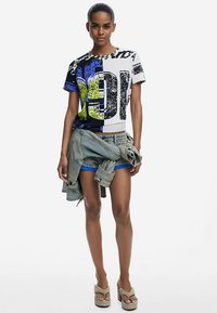 Tricou grafic cu un amestec de culori albastre, negre și albe; prezintă text galben îndrăzneț și modele abstracte. Asortat cu pantaloni scurți din denim și sandale.