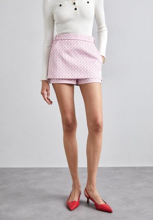 Kvinde iført hvid ribbet langærmet top, lyserød mønstret skort og røde spidse slingback-hæle, der står på gråt gulv.