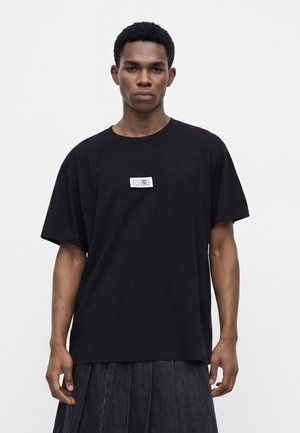 Ung mand med kort krøllet hår iført en løs, sort T-shirt med et lille hvidt mærke på brystet og mørke, plisserede bukser.