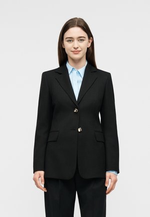 Jeune femme aux cheveux bruns raides, portant un blazer noir texturé sur une chemise bleue claire à col, debout devant un fond uni clair.