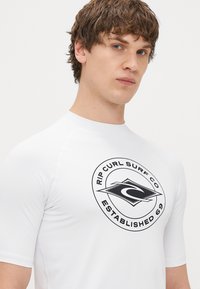 Biały rash guard z krótkim rękawem, z dużym czarnym okrągłym logo z napisem "RIP CURL SURF CO ESTABLISHED 69" wyraźnie wyeksponowanym.