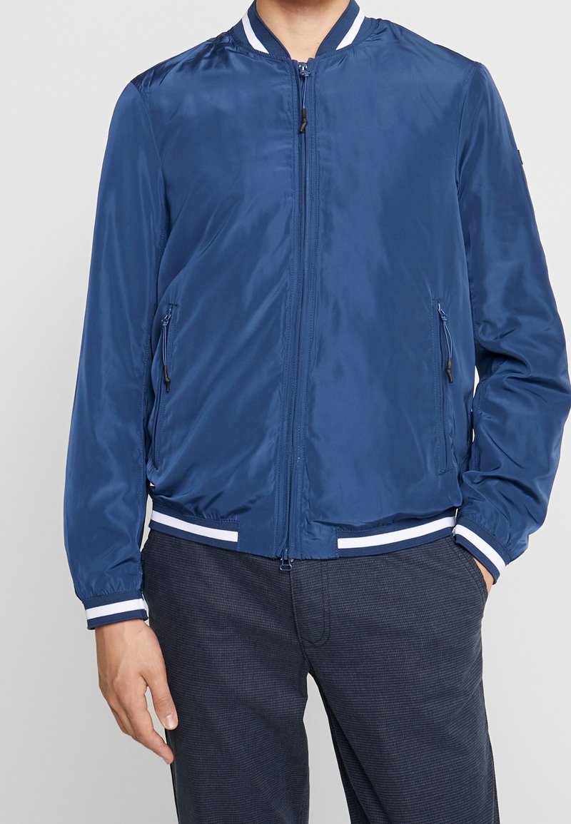 Homme portant une veste bomber bleu marine avec des poignets et un col rayés blancs, associée à un pantalon à carreaux foncé, debout les mains dans les poches.