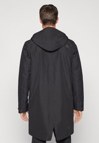 Juodas gobtuvu paltas ilgu kirpimu, turintis tekstūruotą paviršių, šonines kišenes ir logotipą viršutinėje nugaros dalyje.