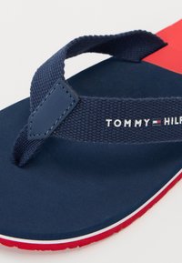 Marine flip-flop med en teksturert stoffrem og gummisåle. "TOMMY HILFIGER" logo trykt på remmen; røde og hvite detaljer på sålen.