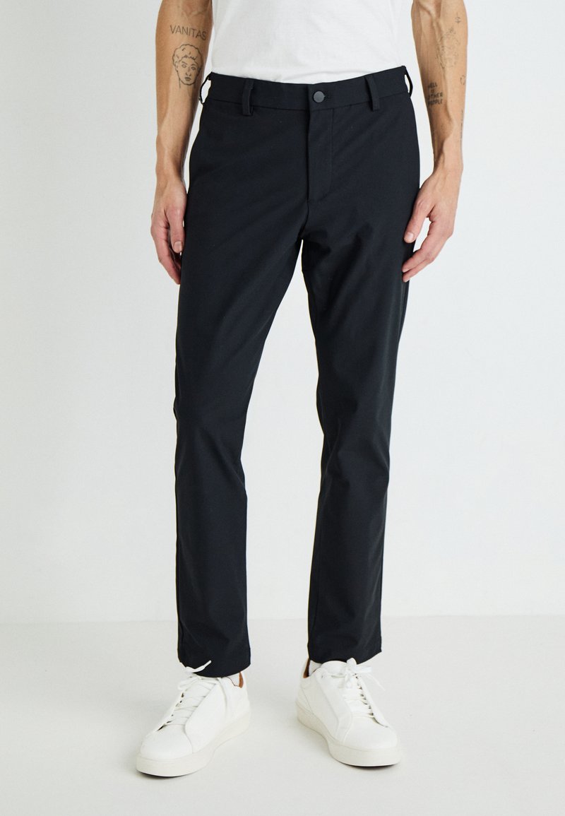 Pantalon noir ajusté au design classique, doté d'une fermeture à bouton, de passants de ceinture et d'un tissu lisse et léger. Associé à des baskets blanches.
