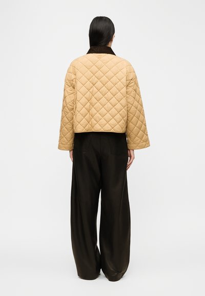 Γυναίκα με ίσια σκουρόχρωμα μαλλιά που φοράει μπεζ, καπιτονέ μπουφάν με φαρδιά μανίκια και μαύρο παντελόνι φαρδιάς γραμμής, στεκόμενη με την πλάτη προς τα εμάς.