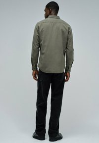 Hombre de espaldas vestido con una camisa de mangas largas verde oliva, jeans negros y zapatos negros frente a un fondo gris liso.