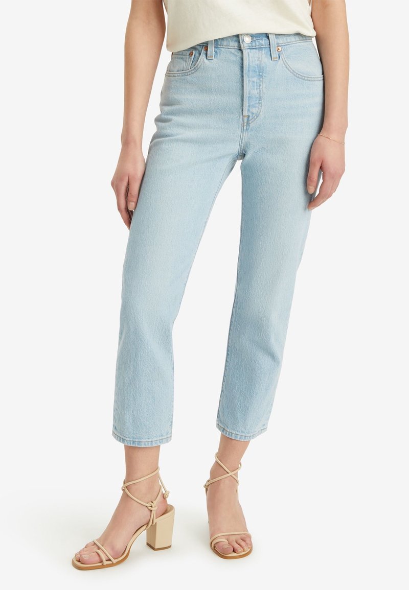 Femme portant un jean court bleu clair taille haute et des sandales beige à talons blocs et brides, debout devant un fond blanc.