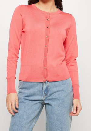 Frau trägt eine korallrosa, geknöpfte Strickjacke mit langen Ärmeln und hellblaue, hoch taillierte Jeans, steht vor einem weißen Hintergrund.