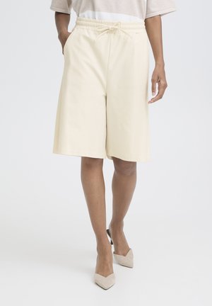 Kvinde iført løse, knælange cremefarvede shorts med elastik i taljen og snøre, kombineret med beige spidse hæle.