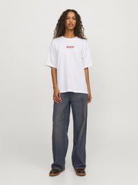 Camiseta de algodón blanca con el texto rojo "HOWDY", combinada con unos pantalones vaqueros oscuros de corte holgado y zapatos marrones con una suela texturizada.