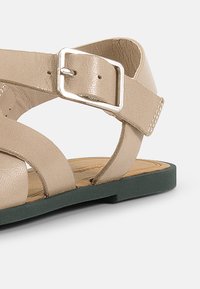 Beige nahka-sandaalit ristikkäisillä hihnoilla ja suorakulmaisella hopeisella soljella. Varustettu tekstuurilla varustetulla pohjalla ja tummanvihreällä litteällä pohjalla.