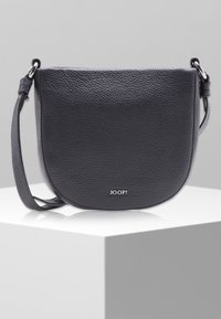 Czarna, teksturowana skórzana torba na ramię z zaokrąglonym dnem i regulowanym paskiem, leżąca na białym stoliku wystawowym na szarym tle.