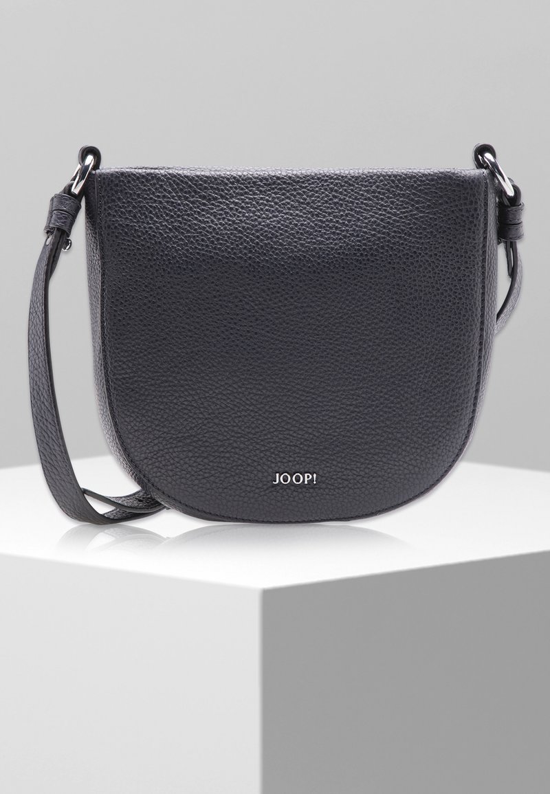 Czarna, teksturowana skórzana torba na ramię z zaokrąglonym dnem i regulowanym paskiem, leżąca na białym stoliku wystawowym na szarym tle.