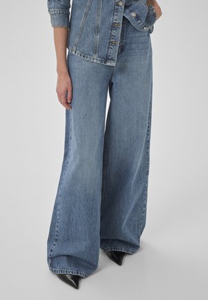 Persona che indossa jeans larghi in denim blu e una giacca in denim abbinata con scarpe nere appuntite su uno sfondo neutro.