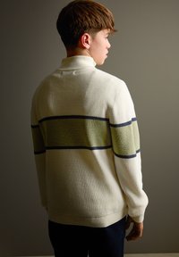 Pull en maille couleur crème avec une finition texturée, présentant des rayures horizontales en olive et bleu marine. Col montant et coupe décontractée.