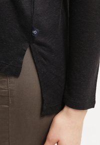 Schwarzes langärmliges Oberteil mit einem seitlichen Schlitz, aus strukturiertem Stoff, mit einem kleinen navy-farbenen Etikett am Saum. Getragen über hellbraunen Hosen.