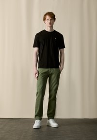 Zwarte T-shirt met korte mouwen en klein logo, gecombineerd met groene broek. Model draagt witte sneakers. Neutrale achtergrond.