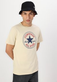 Camiseta de algodón beige con un logo redondo que presenta una estrella azul y el texto "Converse All Star" en rojo, combinada con un sombrero tipo bucket negro.