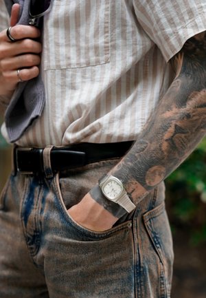 Persona con camisa de rayas y jeans de lavado ácido, con cinturón negro, reloj de pulsera plateado y brazo tatuado, mano en el bolsillo.