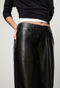 Čierne nohavice typu wide-leg z umelého koženého materiálu s hladkým povrchom, prednými vreckami a bezpečným pásom. Prispôsobený dizajn s čistými švami.