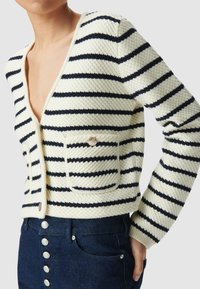 Cardigan a righe realizzato in tessuto testurizzato di colore crema con strisce blu navy, dotato di scollatura a V profonda, due tasche frontali e bottoni decorativi.
