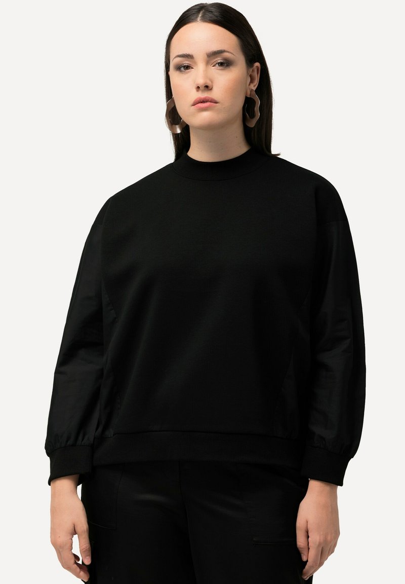 Sudadera negra con cuello alto y mangas largas, ligeramente abullonadas. La tela parece suave y ligeramente texturizada, con un corte relajado.